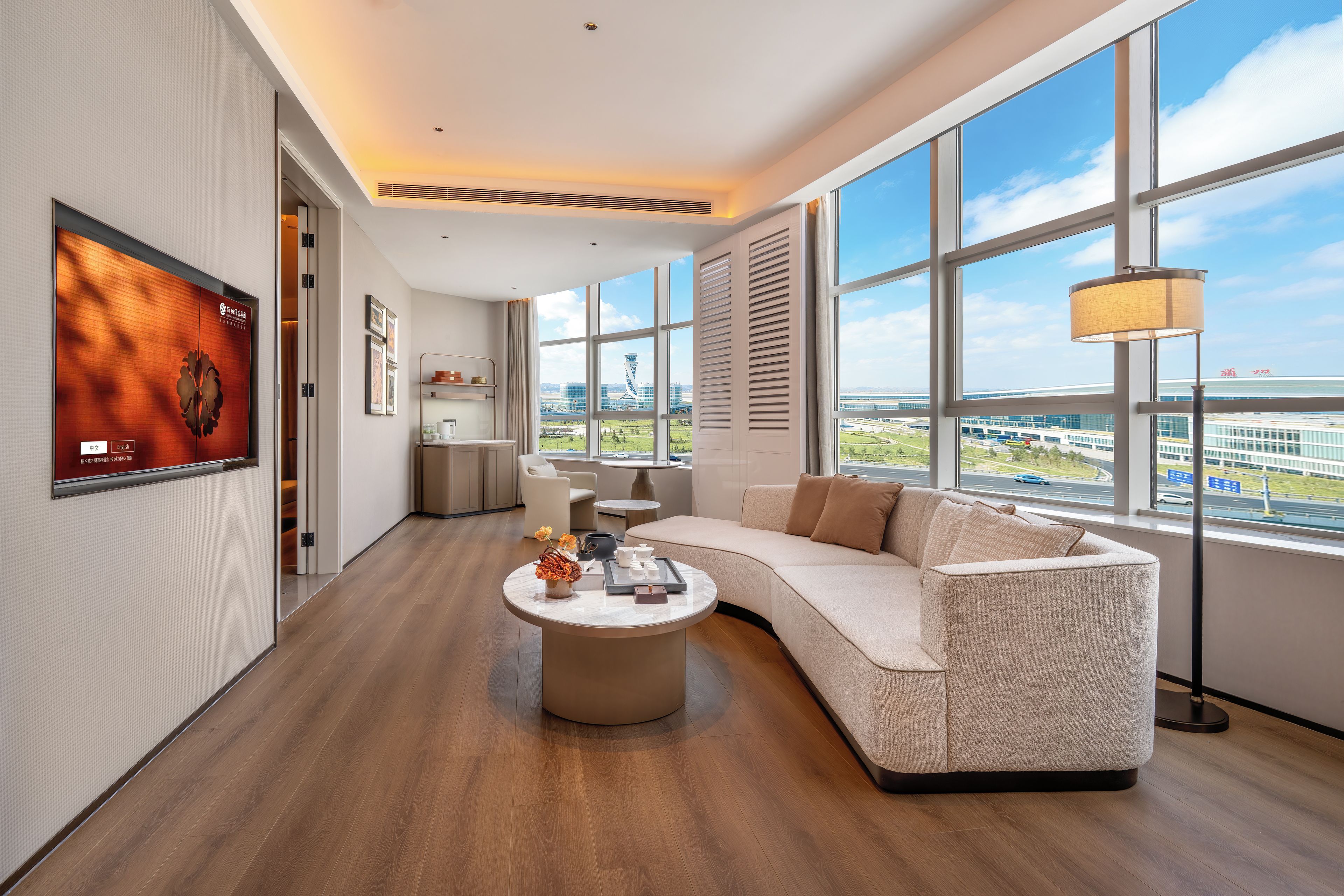 A modern living room with a large window, a sofa, a coffee table, and a TV mounted on the wall.