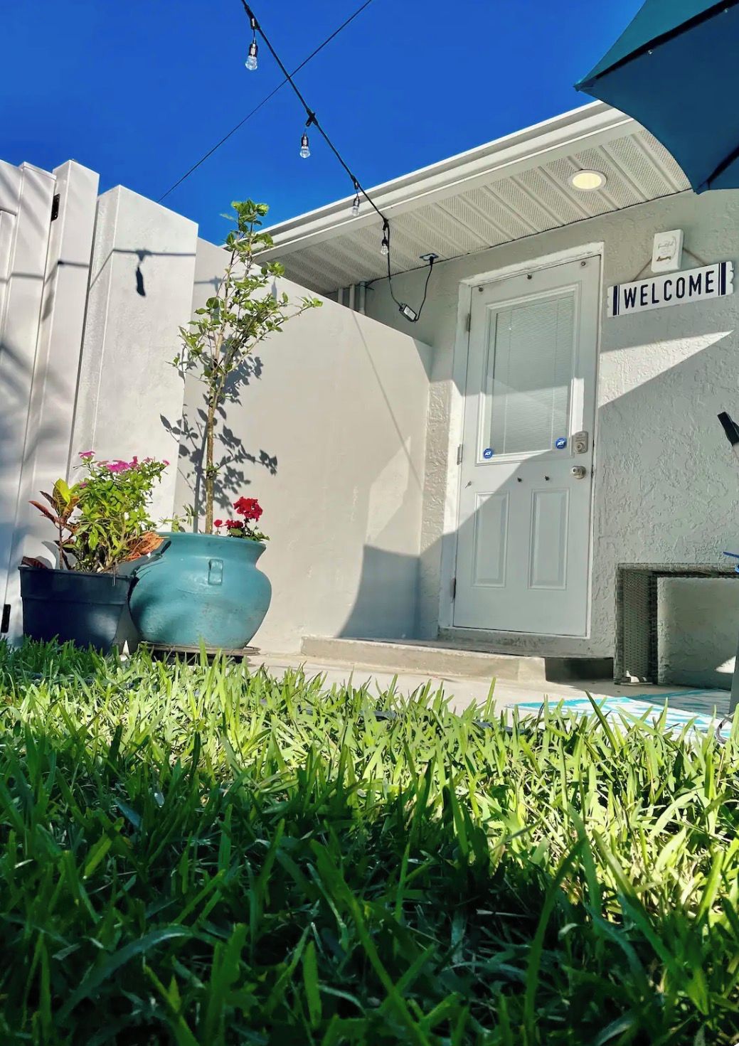 A house with a white exterior, a welcome sign, a door, and a small garden with grass and potted plants.
