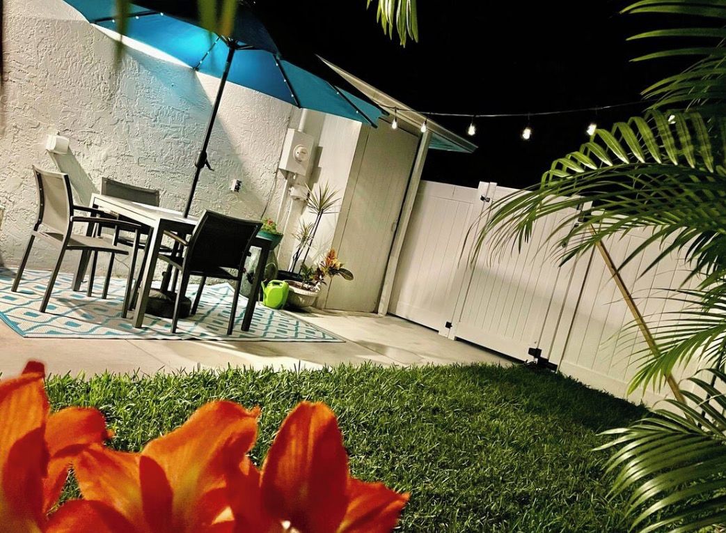 An outdoor patio area with a table and chairs, surrounded by potted plants and a grassy lawn.