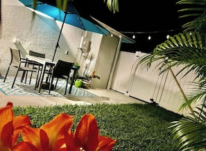 An outdoor patio area with a table and chairs, surrounded by potted plants and a grassy lawn.