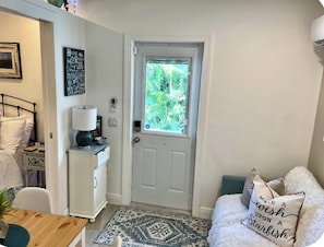 A room with a white door, a small table with a lamp, a white chair, a white sofa with a cushion, and a wall with a framed picture and a sign. - Sweet Suite waiting for you. (Lehigh Acres)