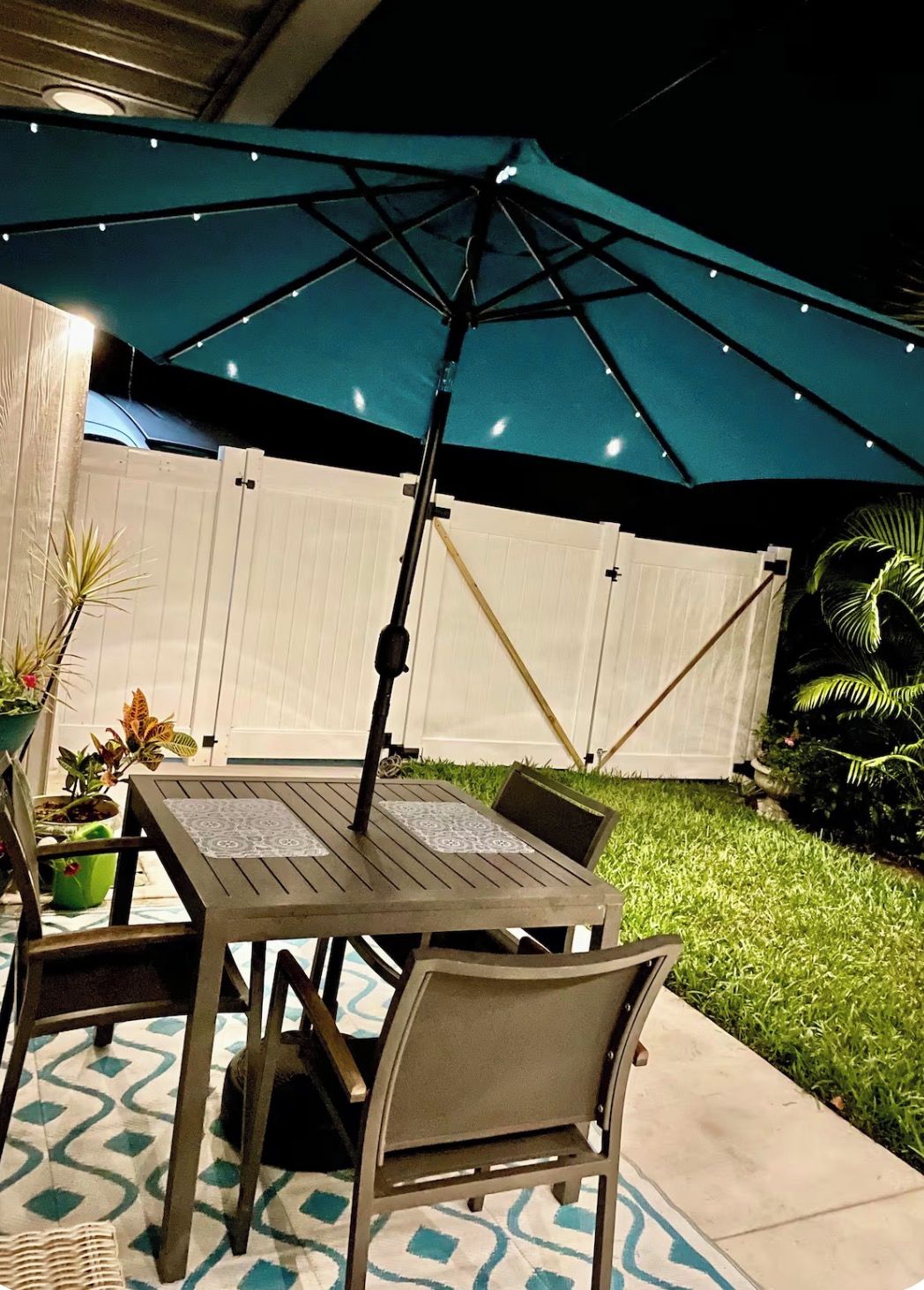 Een terras buiten met een tafel, stoelen, een blauwe parasol en potplanten.