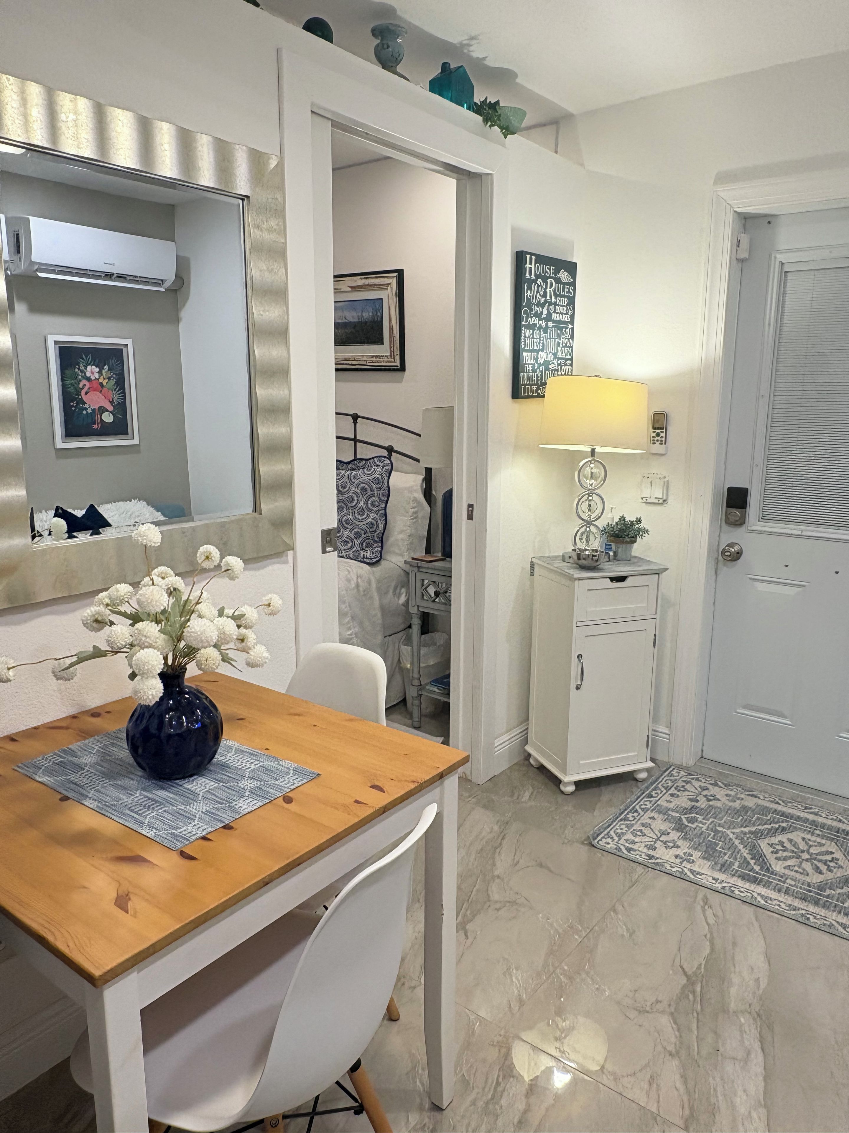 A room with a wooden dining table, a white chair, a vase with white flowers, a mirror, a framed picture, a bedside table with a lamp, a door, and a small cabinet.
