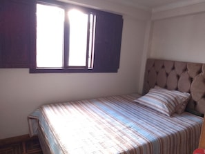 A single bed with a striped bedspread and a headboard, a window with curtains, and a small bedside table. - The Awakening of the Inca (Cusco)