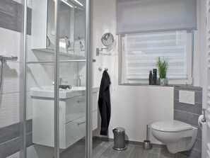 A modern bathroom with a glass shower enclosure, a white toilet, and a window with blinds. - Holiday Apartment With 1 Bedroom (St. Peter-Ording)