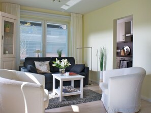 Een woonkamer met een bank, twee fauteuils, een salontafel en een vloerlamp. Door een open deur is een keuken te zien met een fornuis en kasten.