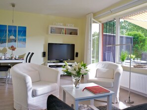 A living room with a television, two white armchairs, a coffee table with a book and flowers, and a bookshelf with books and decorative items. - Holiday Apartment With 1 Bedroom (St. Peter-Ording)