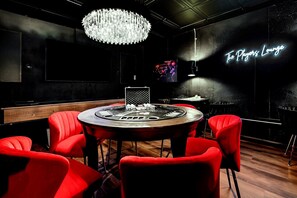 Une pièce avec une table ronde, des chaises rouges, un lustre et un panneau néon sur lequel on peut lire : « The Players Lounge ».