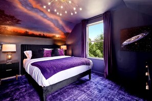 Une chambre à coucher avec un lit, des tables de chevet, une fenêtre et une fresque murale sur le thème de la galaxie.