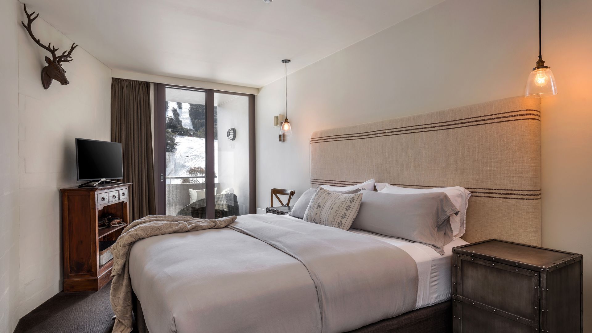 Une chambre à coucher avec un lit, un téléviseur posé sur un meuble en bois, une fenêtre avec des rideaux et une décoration murale représentant une tête de cerf.