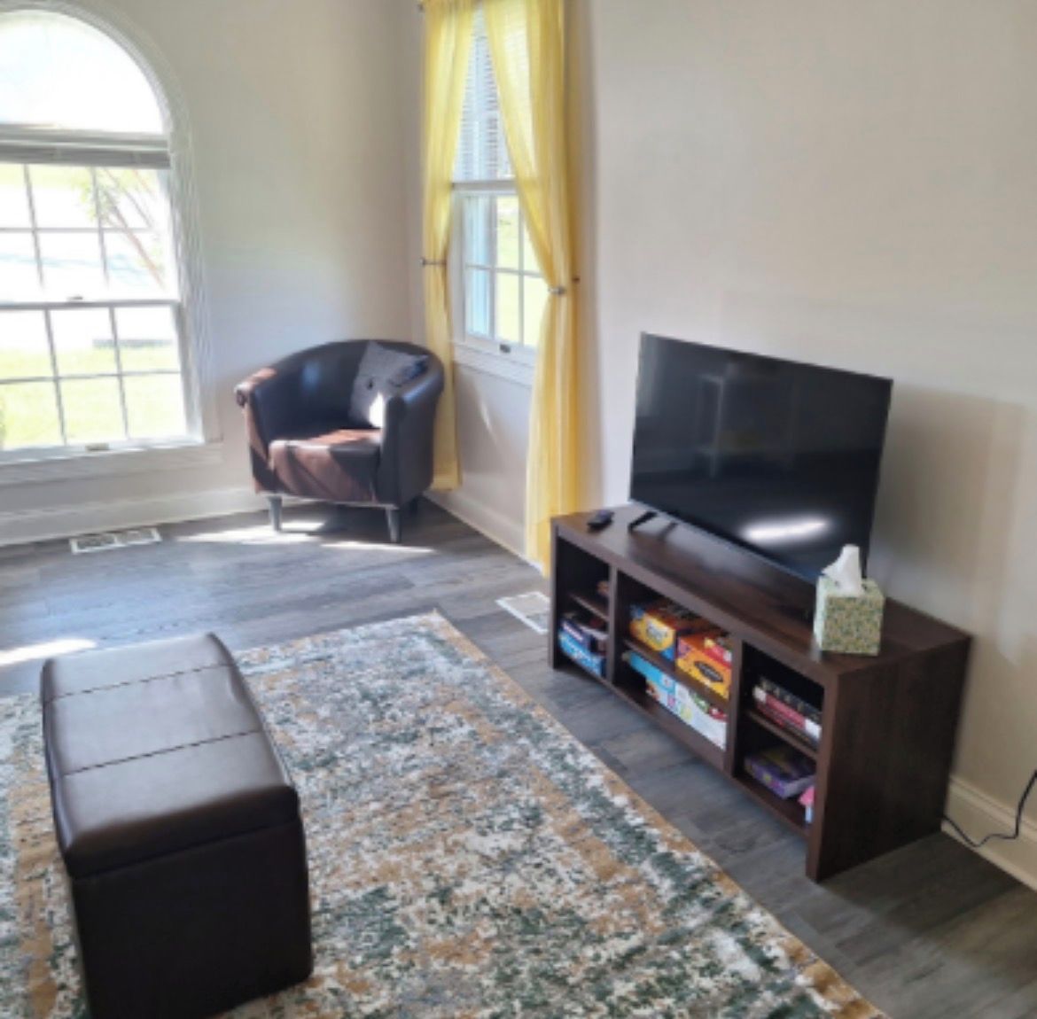 Una sala de estar con una televisión, un sillón de cuero, un tapete y una ventana con cortinas.