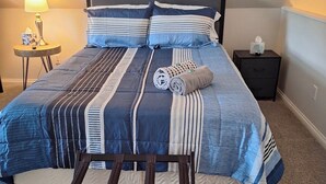 En pænt redt seng med stribet sengetøj, et sengebord med en lampe, og et lille natbord med en tissueske.