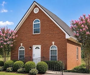 A brick building with a gabled roof, a white door, and two windows with white frames.