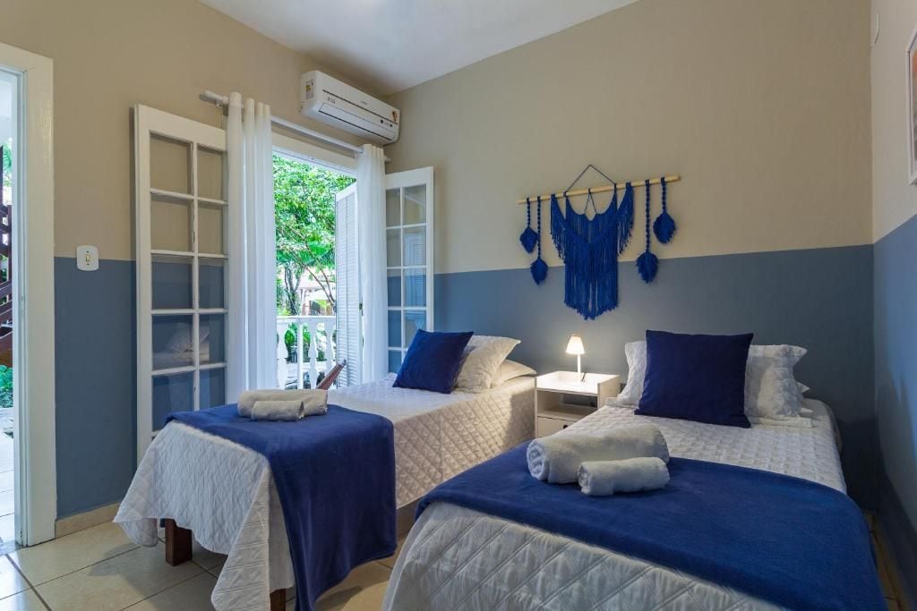 Quarto com duas camas, janela com cortinas, ar-condicionado e decoração de macramé na parede.