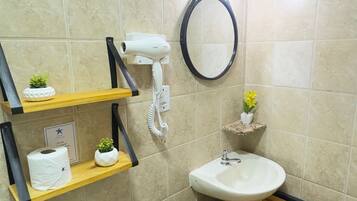 Un baño con inodoro, lavamanos y espejo. Hay repisas con artículos de aseo personal y una planta pequeña.