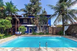 Una piscina con borde de piedra, rodeada de vegetación tropical y un edificio de dos pisos con fachada azul.