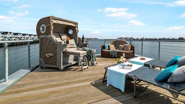 A deck with a wicker sofa, a lounge chair, and a small table with a bottle and flowers. - Houseboat in Olpenitz With Swimming Pool (Kappeln)