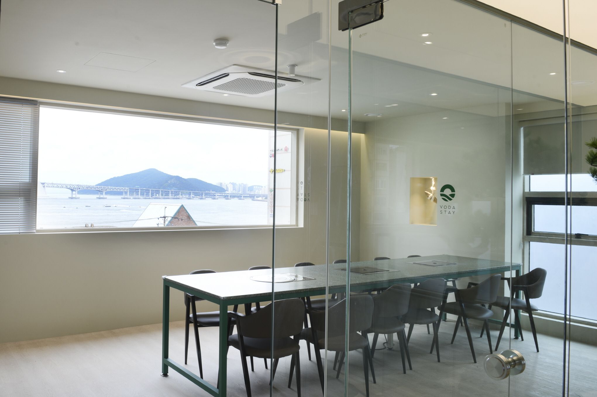 Ein Konferenzraum mit Glastisch und Stühlen, Blick auf eine Brücke und Berge durch das Fenster sowie ein 'Voda Stay'-Logo an der Wand.