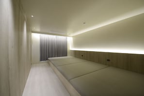 Ein Zimmer mit einem langen, schmalen Bett, einem Fenster mit Vorhängen und einer Wandleuchte.