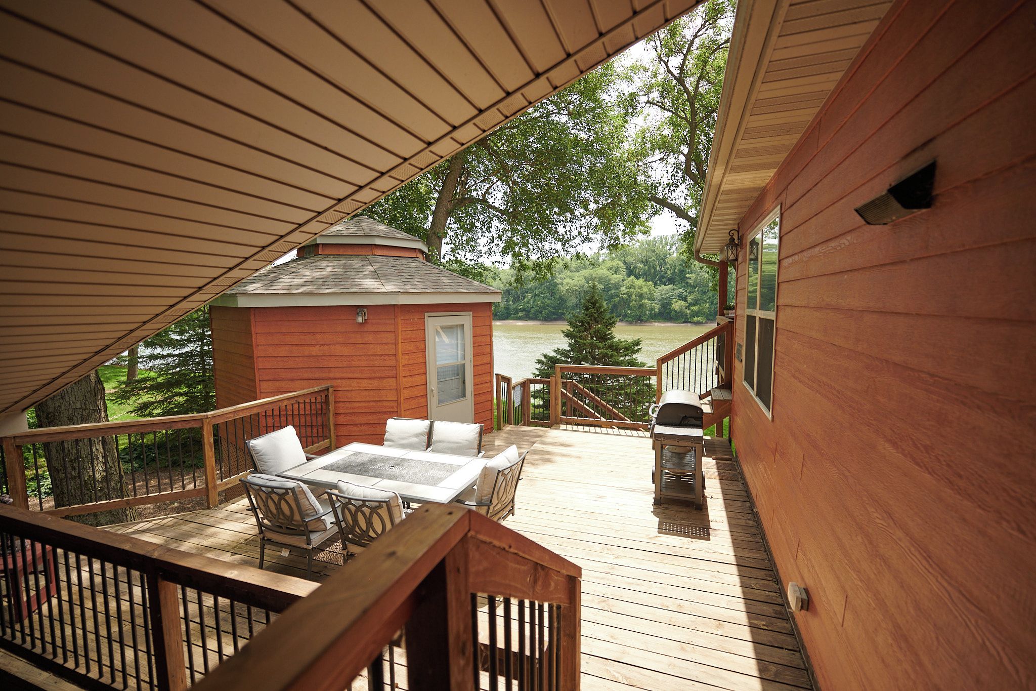 Una terraza de madera con mobiliario exterior, una cabaña pequeña y vistas a un lago.