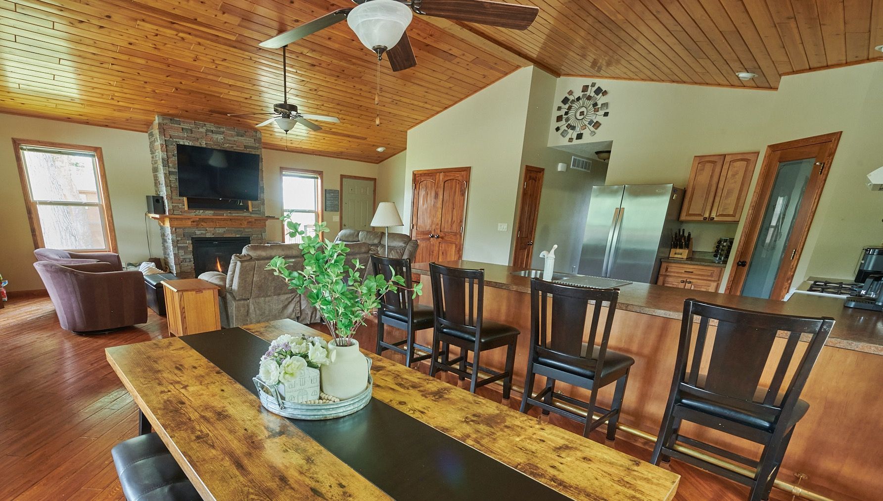 A spacious living area with a wooden ceiling, a fireplace, a dining table with chairs, and a kitchen area with cabinets and appliances.