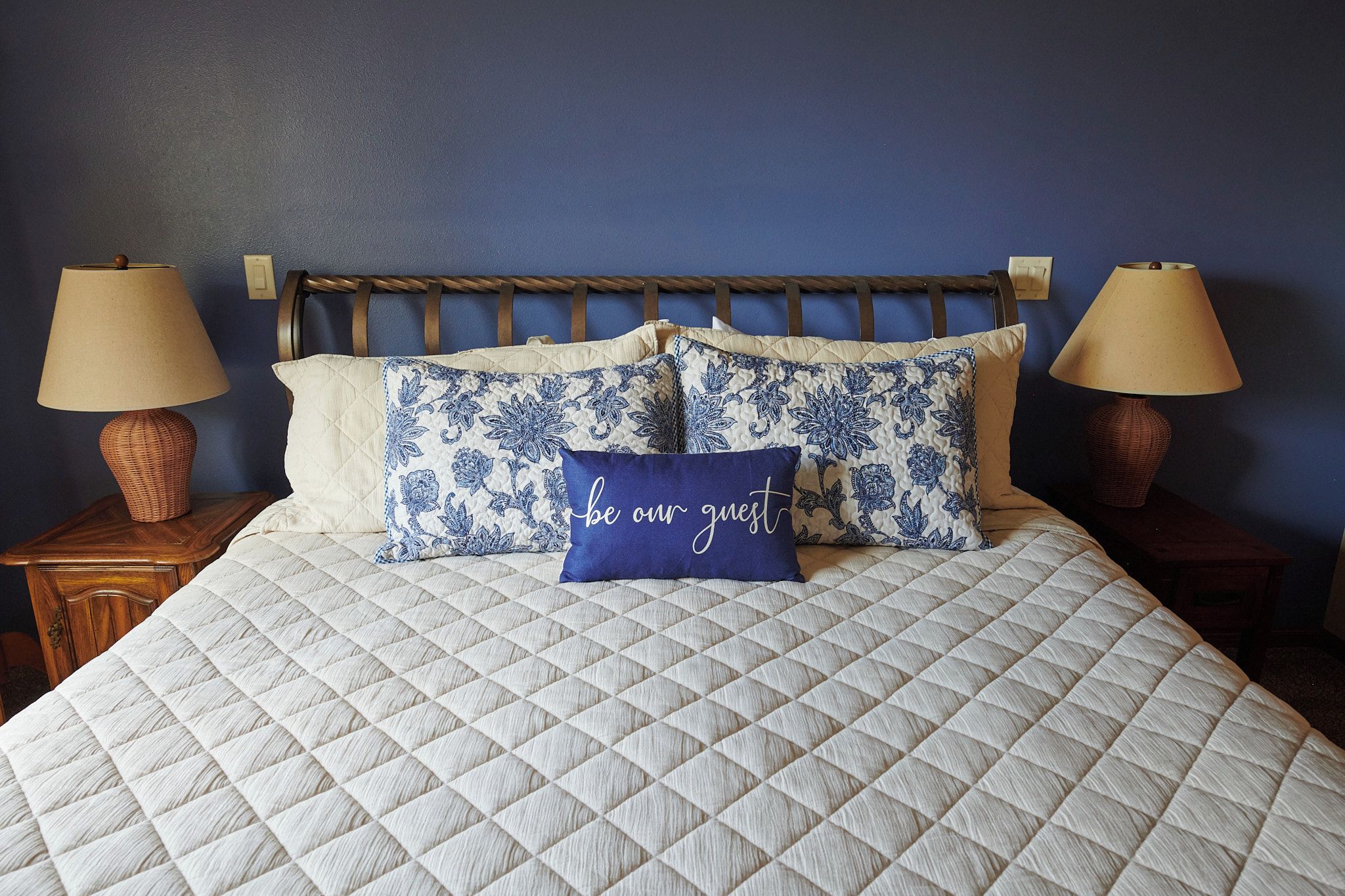 Un letto con copripiumino trapuntato, due abat-jour e cuscini decorativi con motivi floreali, più un cuscino blu con scritte.