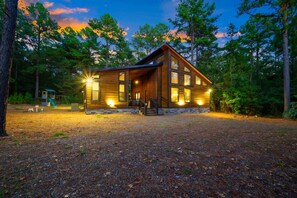 A two-story wooden cabin with a covered porch and a small play area in the background. - Luna Wolf Lodge · New Listing! Hot Tub, Fire Pit & Loft Bunks in the Pines (Broken Bow)