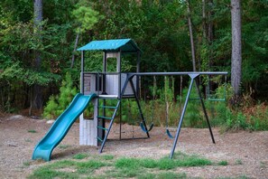 A playground with a slide and swings, surrounded by trees. - Luna Wolf Lodge · New Listing! Hot Tub, Fire Pit & Loft Bunks in the Pines (Broken Bow)