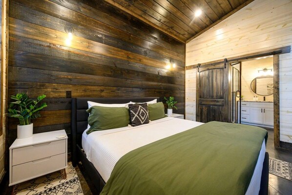 Un dormitorio con cabecera de madera, cama con ropa de cama verde, mesita de noche con una planta y un baño visible a través de una puerta abierta.