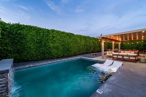 Ein Schwimmbecken mit Wasserfall, umgeben von einer Hecke, und ein Außenbereich mit Sitzgelegenheiten und einer Pergola.