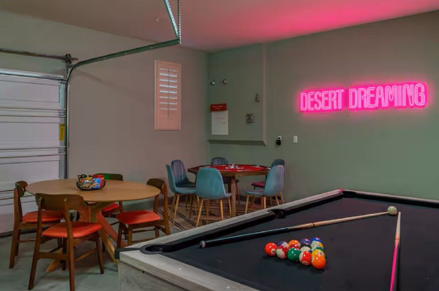 A pool table with billiard balls, a neon sign reading 'DESERT DREAMING,' and a dining area with chairs and a table.