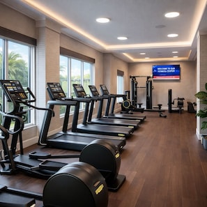 Ein Fitnessraum mit Laufbändern, Crosstrainern und einem Fernseher, der 'WILLKOMMEN IN UNSEREM FITNESSRAUM' anzeigt.