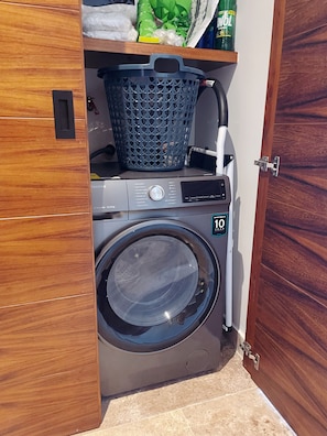 A compact laundry area with a washing machine and dryer, a basket, and a shelf with items. - 5th Ave. Studio Ocean View Rooftop (Playa del Carmen)