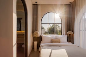 A bedroom with a large arched window, two bedside lamps, a bed with white linens, and a view of palm trees outside. - NEW 3BR Boutique Villa w/ Private Pool - Bingin (South Kuta)