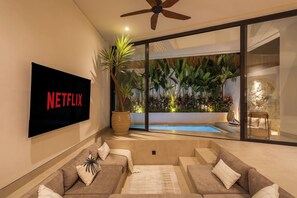 Een moderne woonkamer met een flatscreen televisie waarop het Netflix-logo te zien is, een grote potplant, een zwembad met glazen wanden en een bank met kussens.