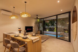 A modern dining area with a TV displaying 'NETFLIX,' a pool visible through a glass wall, and a ceiling fan. - Belluci Uluwatu | 1BR Modern Villa w/ Pvt Pool (South Kuta)