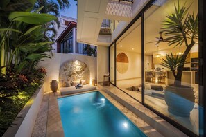 A modern outdoor pool area with a stone wall, seating, and a view into a well-lit interior space. - Belluci Uluwatu | 1BR Modern Villa w/ Pvt Pool (South Kuta)