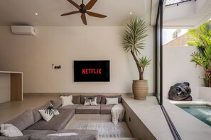 A modern living room with a sectional sofa, a ceiling fan, a large potted plant, a wall-mounted TV, and a view of a swimming pool. - Belluci Uluwatu | 1BR Modern Villa w/ Pvt Pool (South Kuta)