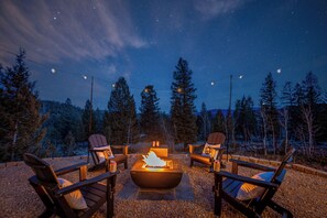 A night scene with a fire pit surrounded by Adirondack chairs, set against a backdrop of a starry sky and tall trees. - Modern Private 3 Acres w/ Hot Tub and Game Room (Bailey)