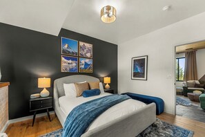 A modern bedroom with a large bed, two bedside tables, a wall-mounted lamp, and a framed picture of a mountain landscape. - Modern Private 3 Acres w/ Hot Tub and Game Room (Bailey)