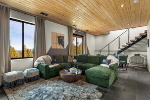A modern living room with green sofas, a coffee table, and a large window with a view of trees. - Modern Private 3 Acres w/ Hot Tub and Game Room (Bailey)