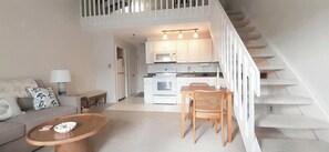 A modern living room with a sofa, coffee table, and a dining area with chairs. A staircase leads to a kitchen with white cabinets and a microwave. - Steps from Wells Beach, Bellevue by the Sea is perfect for your Maine vacation (Wells)