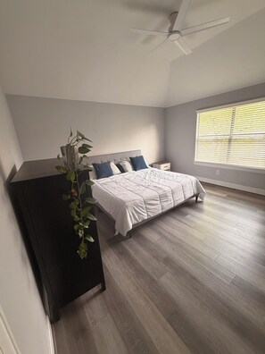 Ein Schlafzimmer mit einem Bett, einem Nachttisch, einem Deckenventilator, einem Fenster mit Jalousien und einer Topfpflanze.