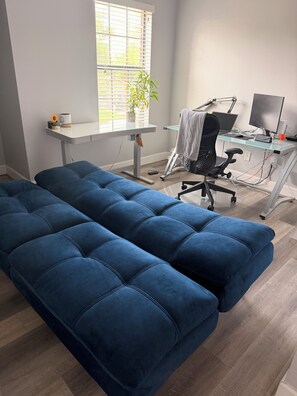 Ein Zimmer mit einem blauen Sofa, einem Schreibtisch mit Computer und einem Fenster mit Jalousien.