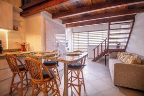 A modern kitchen with a dining area, wooden ceiling beams, and a staircase leading to a living area with a sofa.