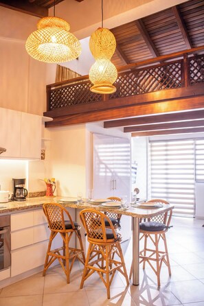 A modern kitchen with a dining area, featuring wicker chairs, pendant lights, and a wooden ceiling.