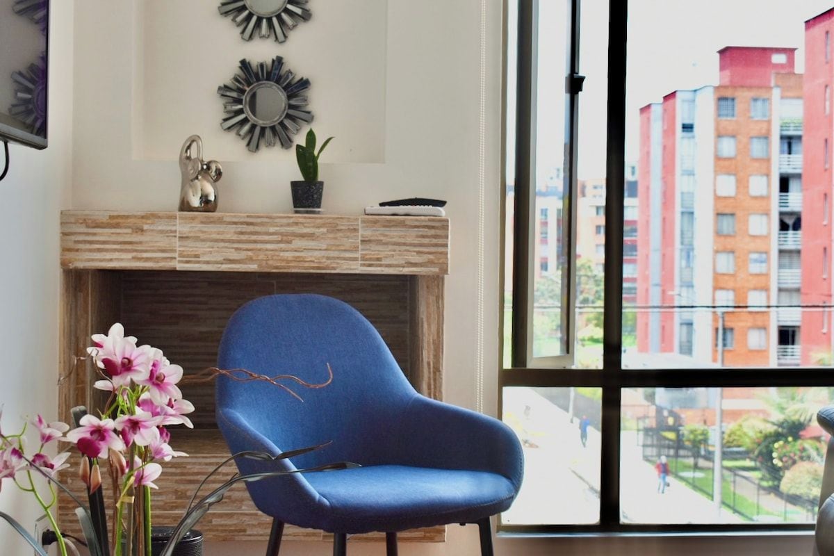 Une pièce avec un fauteuil bleu, une cheminée et une vue sur les bâtiments, visible à travers une grande fenêtre.