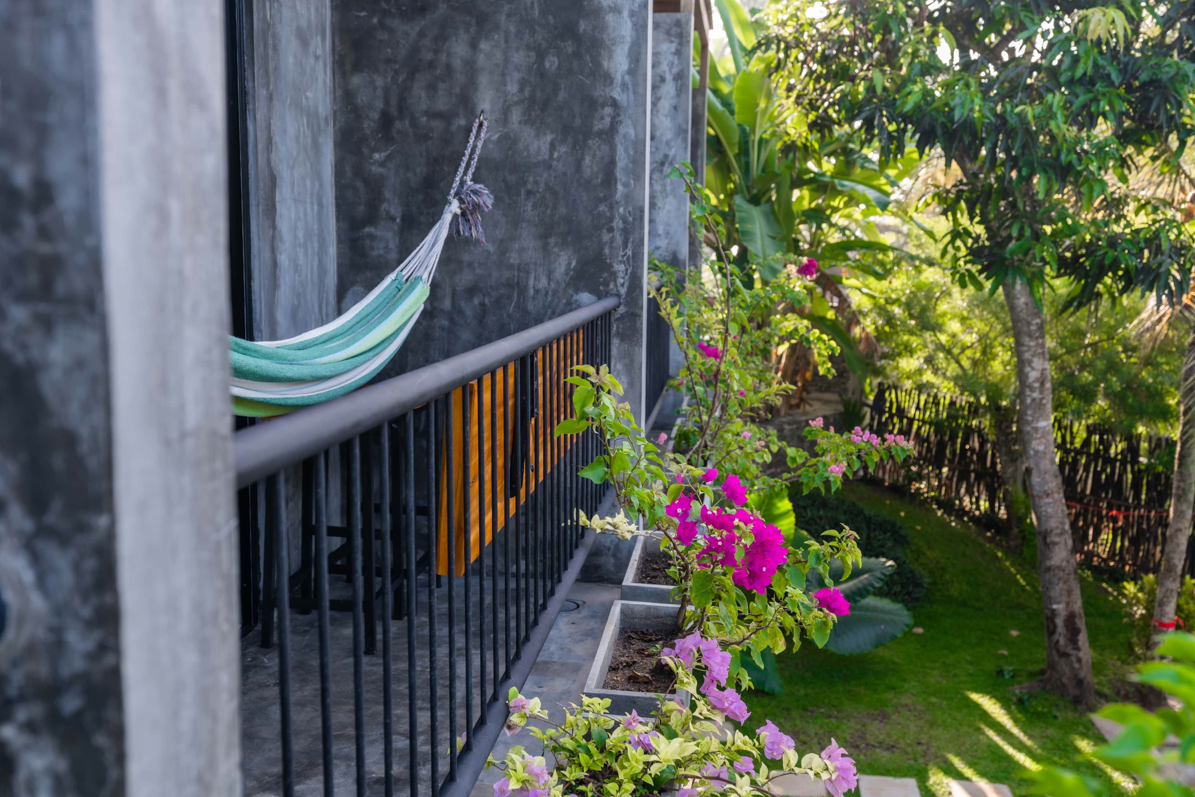 Uma rede pendurada em um corrimão de metal, uma parede de concreto e um jardim com flores cor-de-rosa e folhagem verde.