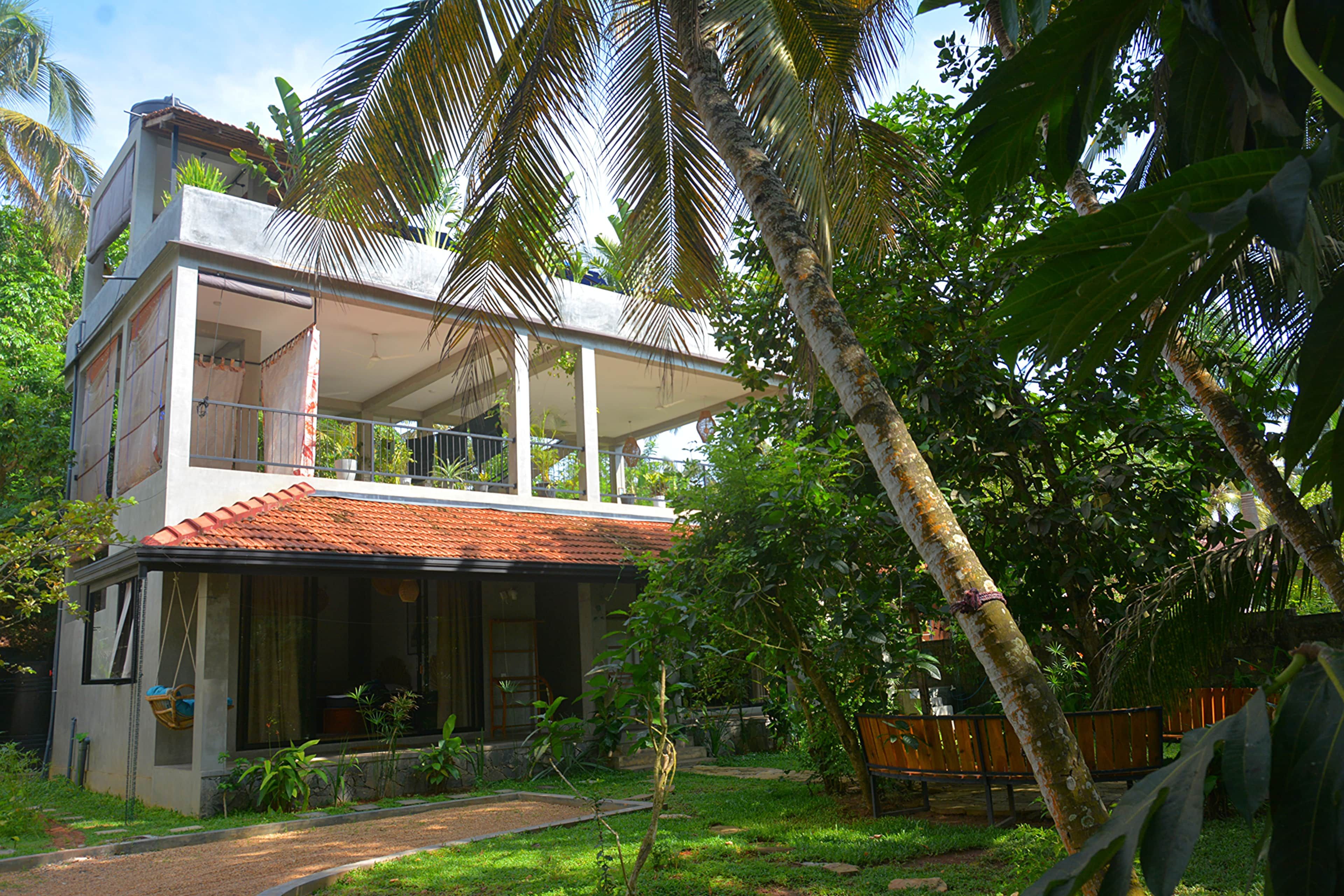A two-story house with a balcony, surrounded by lush greenery and a palm tree.