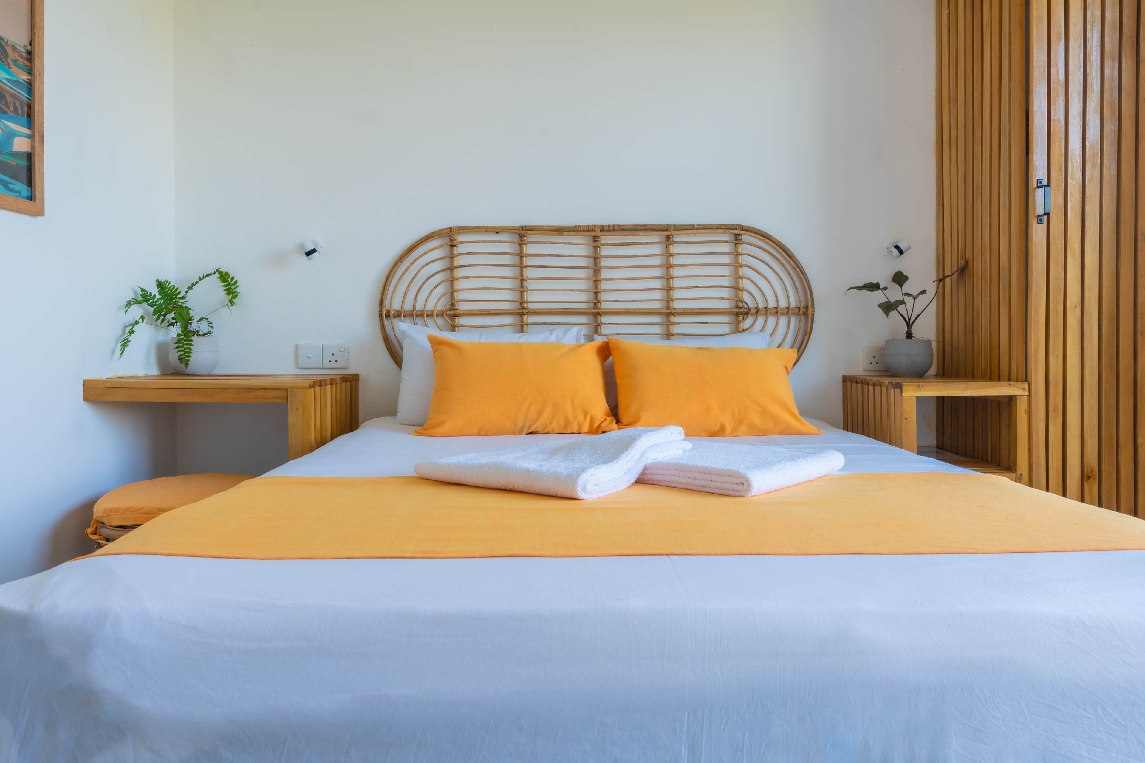 Ein ordentlich bezogenes Bett mit weißer und orangefarbener Bettdecke, zwei Kissen und gefaltete Handtücher. Auf jedem Nachttisch stehen eine Lampe und ein Blumentopf.
